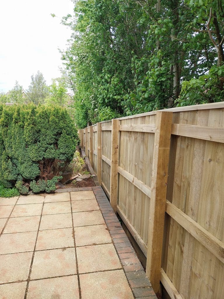 Wooden fence along a garden path with trees and shrubs on the left. The path is flanked by square stone tiles bordered by a brick line on the right.
