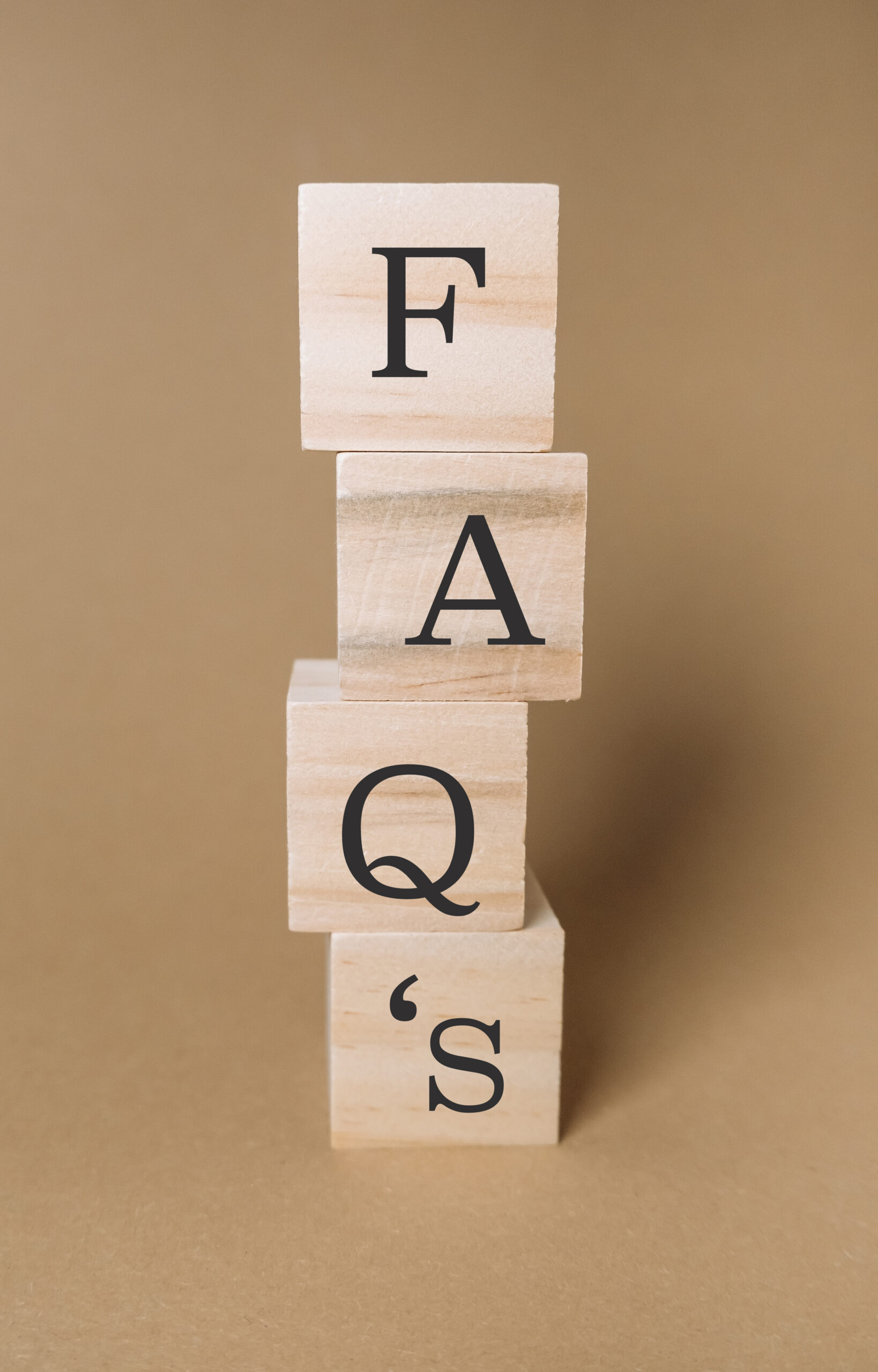 Four wooden blocks stacked vertically, each displaying a black letter to spell "FAQ's" against a plain brown background.