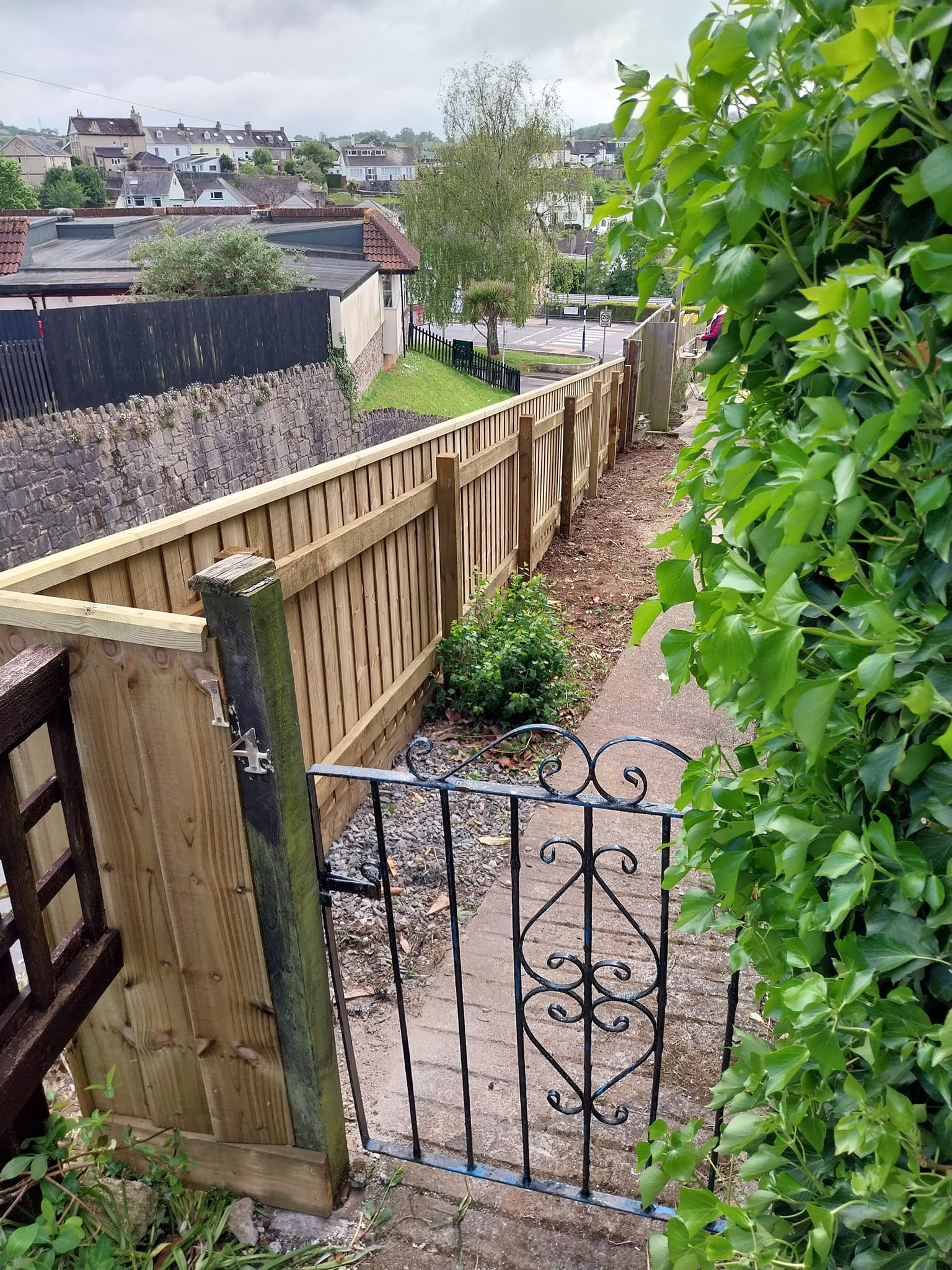 A narrow garden path with a metal gate leads alongside a wooden fence, bordered by green shrubs. Houses and rooftops are visible in the background under a cloudy sky.