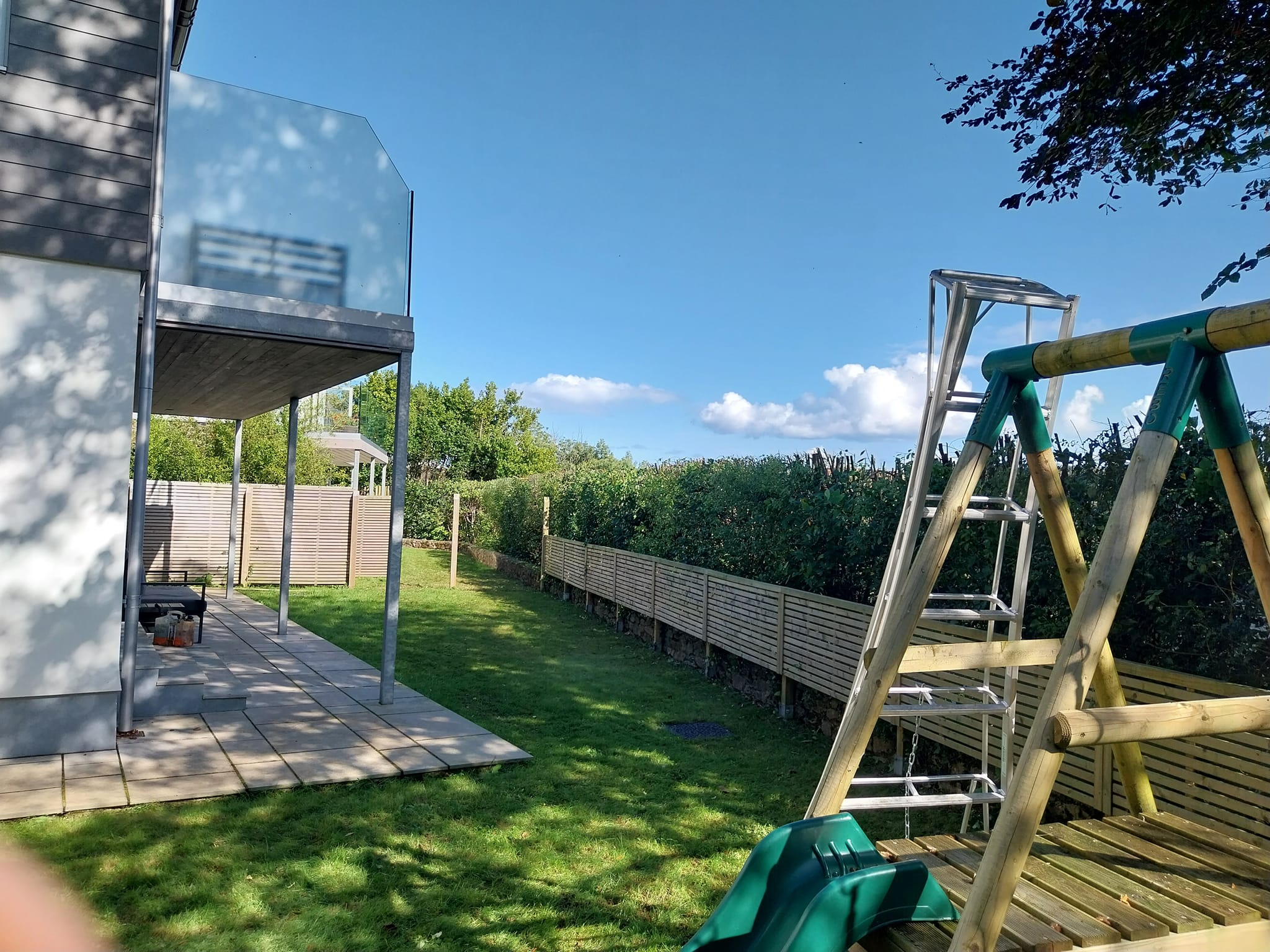 A backyard with a swing set, a metal ladder, and a modern house with a balcony. There is a patio with outdoor furniture, green grass, a wooden fence, and trees under a clear blue sky.